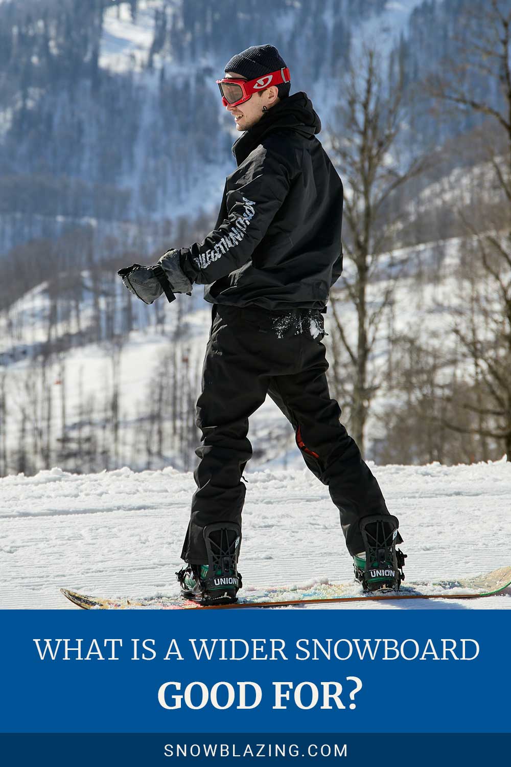 Smiling man in a snowboard track wearing black jacket and red googles - What Is a Wider board good for?
